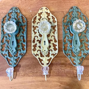 May include: Three wall hooks featuring ornate, openwork metal backplates in teal and cream colours. Each hook includes a clear, faceted glass knob at the top and a smaller, clear glass hook at the bottom, designed for mounting on a flat surface.