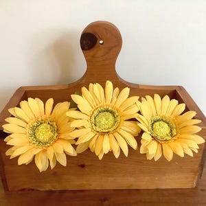 May include: A rectangular wooden caddy with a central handle featuring a circular cutout at the top. Three artificial yellow sunflowers with textured green and yellow centres are arranged horizontally inside the open wooden container.