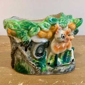 May include: A ceramic planter with a whimsical design. The planter features a deer figure and tree trunk with green leaves, orange fruit, and a gray and white colour scheme. The planter is a decorative item.