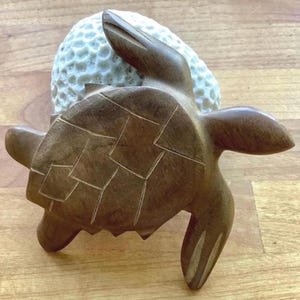 May include: A carved brown wooden sea turtle figurine positioned in front of a white, textured, spherical object with a dimpled surface. The turtle features detailed shell segments and smooth, polished limbs, resting against the round white item.