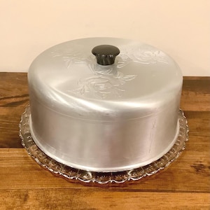 Peut inclure: Un porte-gâteau argenté vintage avec un bouton noir sur le dessus, posé sur une assiette en verre transparent à bords festonnés. Le couvercle présente des motifs floraux en relief. Un article de cuisine classique.