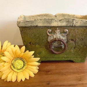 Peut inclure: Une jardinière rectangulaire en céramique vert olive avec un bord crème patiné et un ornement central en forme d'anneau métallique. Deux tournesols artificiels jaunes aux centres pailletés sont placés devant la jardinière.
