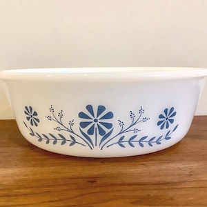 May include: A white round baking dish with integrated side handles, featuring a blue floral pattern around the exterior. The design consists of a central stylised flower flanked by smaller floral sprigs and leaves, all rendered in a solid blue.