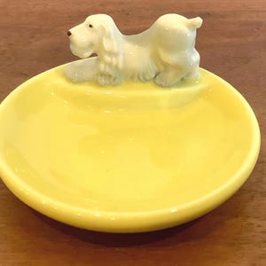 Peut inclure: Un petit plat jaune en céramique avec une figurine de chien blanc. Le plat est rond et brillant. Le chien est couché sur le bord du plat. Idéal pour ranger de petits objets.