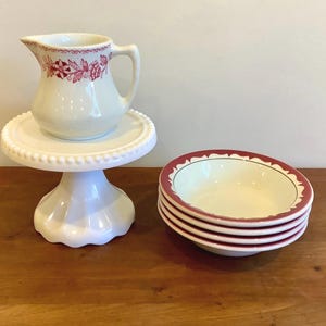 Peut inclure: Un pot à lait en céramique blanche avec un motif floral rouge est posé sur un présentoir à gâteaux blanc à perles. À côté, une pile de quatre bols blancs, chacun doté d'un bord rouge avec un motif festonné sur le pourtour intérieur.