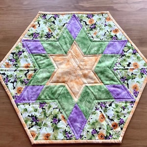 Peut inclure: Un tapis de table matelassé hexagonal présentant un motif d'étoile central dans des tons pêche, vert et violet. La bordure extérieure se compose d'un tissu à motif floral avec des fleurs blanches, orange et violettes sur fond noir, fini par un liseré pêche.