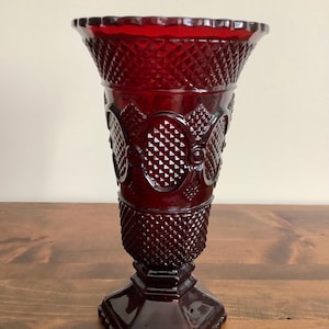 May include: A ruby red glass vase with a diamond pattern and a scalloped rim. The vase has a flared top and a pedestal base. The vase is approximately 25 cm tall.