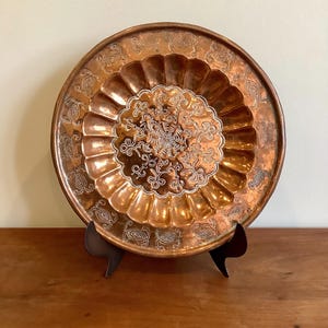May include: A round copper plate featuring a central medallion with intricate etched floral patterns, surrounded by a scalloped border and an outer rim decorated with repeating oval motifs. The metal has a warm, polished finish throughout.