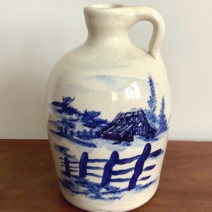 May include: A cream-coloured ceramic jug with a handle, featuring a hand-painted blue landscape scene. The design includes a fence, a building, and trees. The jug has a rounded body and a narrow neck.