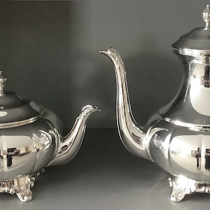 Peut inclure: Une théière et une cafetière assorties en métal argenté poli, présentant toutes deux un corps arrondi à lobes, des anses courbes ornées, un bouton décoratif sur chaque couvercle et quatre petits pieds détaillés sous chaque récipient.