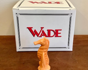 May include: A small, peach-coloured ceramic seahorse figurine standing in front of a white rectangular box. The box features the word WADE printed in bold red capital letters on the front and top, framed by thin black decorative border lines.