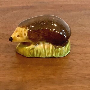 Peut inclure: Une petite figurine de hérisson en céramique avec un dos brun foncé brillant et un visage crème clair doté d'un nez noir. Le hérisson est posé sur une base texturée vert clair imitant un brin d'herbe, présentant une finition lisse.