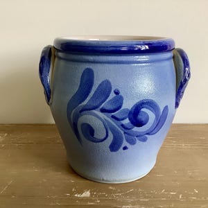 May include: A blue ceramic pot with a dark blue rim and handles. The pot features a lighter blue base colour with a dark blue floral design. The pot is sitting on a wooden surface.