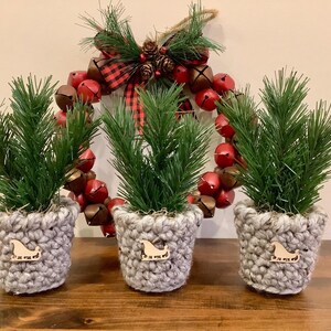 May include: Three small Christmas tree decorations in grey crocheted pots with wooden sleigh accents. A festive wreath with red bells and a plaid bow hangs in the background, creating a holiday theme.
