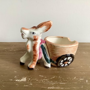 May include: A ceramic donkey planter with a cart. The donkey is white with brown ears and markings, pulling a tan cart with a black wheel. The cart has a white interior, and the donkey has a pink and green harness.