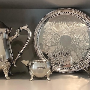 May include: A silver tea set comprising a teapot, serving tray, creamer, and sugar bowl. The set features intricate detailing and is displayed on a shelf. The tray has a decorative border and central design.