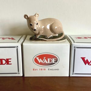 Peut inclure: Figurine Wade en céramique représentant une souris beige sur une base verte, posée sur une boîte blanche. La boîte affiche le logo Wade en rouge et le texte « Est. 1810 England ». Deux autres boîtes avec le logo Wade sont visibles.