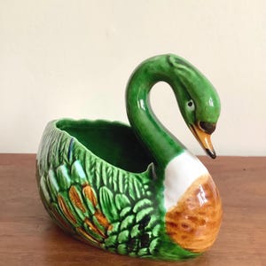 May include: A ceramic swan planter with a green neck and body, white chest, and brown accents. The swan's body is a planter, ideal for small plants or flowers. The swan is on a wooden surface.