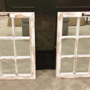 May include: Two rectangular wall mirrors featuring a white distressed wooden frame with a nine-pane window grid design. Each mirror is topped with a dark metal handle, creating a look that mimics a classic window frame with a weathered finish.