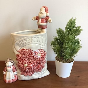 May include: A ceramic planter shaped like a sack with a Santa Claus figure on the rim and a small figure in a white apron at the base. The front features embossed red poinsettia flowers and the text POINSETTIA and GARDEN SEED. Beside it is a small artificial green pine tree in a white pot.