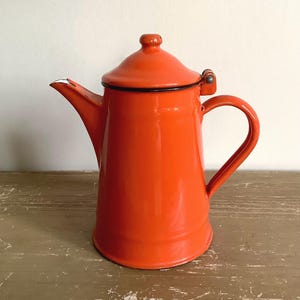 May include: A bright orange enamel coffee pot with a matching lid, a curved handle, and a tapered spout. The pot features a smooth, glossy finish with a dark rim around the lid and base, showing minor signs of wear at the spout tip.