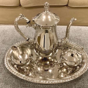 May include: A polished silver tea service set consisting of a tall teapot with a lid, a creamer, and a sugar bowl with a lid, all arranged on an oval silver tray. Each piece features ornate, decorative handles and feet with intricate detailing.