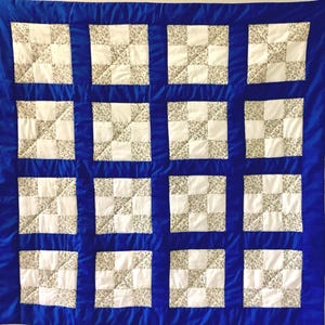 Peut inclure: Une courtepointe carrée composée d'une grille de seize blocs, chacun présentant un motif à neuf carrés alternant entre des carrés blancs unis et des carrés à petits motifs floraux dans des tons sourds, le tout bordé d'un tissu bleu roi.