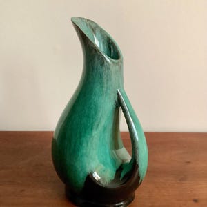 May include: A tall, green and black ceramic jug with a distinctive, curved handle and spout. The jug has a glossy finish with a blend of green and black hues. The design is a modern take on a classic vase.