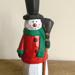 May include: A carved wooden snowman figure. The snowman is wearing a black top hat, a red coat with black buttons, and a green scarf. It has a carrot nose, rosy cheeks, and a black broom.