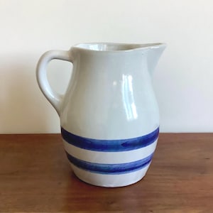 May include: White ceramic jug with a curved handle and spout. The jug has two horizontal blue stripes around the body. The jug is sitting on a wooden surface.