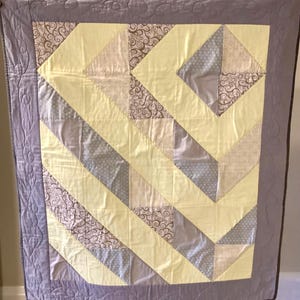 May include: A rectangular quilt featuring a geometric pattern of yellow, grey, and brown swirled fabric blocks. The quilt is bordered by a solid grey frame with a continuous stitched swirl design throughout the border and across the quilt surface.