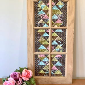 May include: A wooden framed panel with a patchwork design. The patchwork features triangles in shades of blue, pink, green, and brown, set against a dark patterned background. A bouquet of pink and white flowers sits in front of the panel.