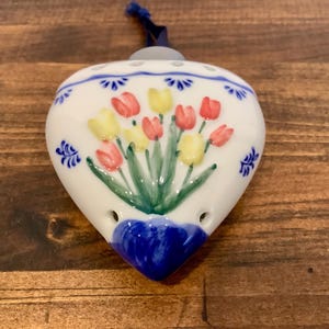 May include: White heart-shaped ceramic ornament with a hand-painted floral design featuring red and yellow tulips. The ornament has a blue ribbon for hanging and is decorated with blue accents. The bottom is a deep blue colour.