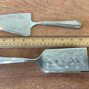May include: Two silver serving utensils with floral patterns. One is a pie server, the other a rectangular cake server. A wooden ruler, approximately 30 cm long, is positioned between them on a wooden surface.