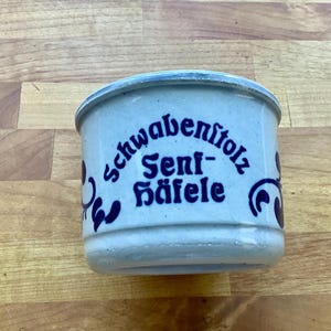 May include: A white ceramic mustard jar with blue lettering that reads "Schwabenstolz Senf-Häfele".