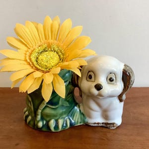 Peut inclure: Un cache-pot en céramique en forme de chiot blanc aux oreilles brunes à côté d'une base verte feuillue, contenant une grande fleur artificielle de gerbera jaune. Le chiot a une finition brillante, des yeux sombres et un petit nez noir.