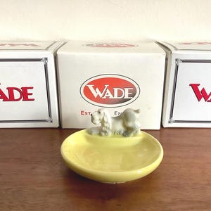 May include: A yellow ceramic dish with a reclining dog figurine. The dish is round and has a shallow bowl shape. The dog is light grey and white. The boxes behind the dish have the word "WADE" in red.