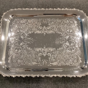 May include: A rectangular silver serving tray featuring ornate, embossed scrollwork and floral patterns across its surface. The tray includes two decorative, scrolled handles on opposite sides and a detailed, scalloped edge border.