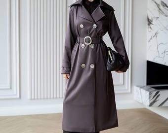 May include: A long, dark gray double-breasted trench coat featuring a wide collar, long sleeves, and a matching belt with a circular buckle. The coat is adorned with metallic buttons and is paired with dark tights, dark heels, and a black handbag.