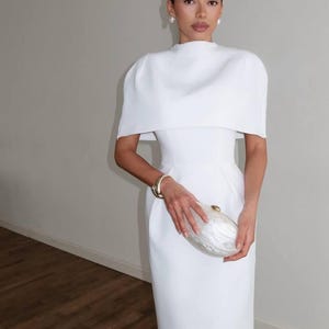 K&ouml;nnte beinhalten: Ein wei&szlig;es, bodenlanges Kleid mit einem einzigartigen, kapuzenartigen Oberteil. Das Kleid hat ein figurbetontes Oberteil und einen stromlinienf&ouml;rmigen Rock. Ein goldenes Armband und eine perlmuttfarbene Clutch vervollst&auml;ndigen den Look.