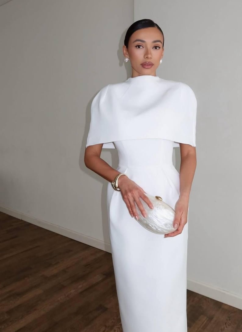 K&ouml;nnte beinhalten: Ein wei&szlig;es, bodenlanges Kleid mit einem einzigartigen, kapuzenartigen Oberteil. Das Kleid hat ein figurbetontes Oberteil und einen stromlinienf&ouml;rmigen Rock. Ein goldenes Armband und eine perlmuttfarbene Clutch vervollst&auml;ndigen den Look.