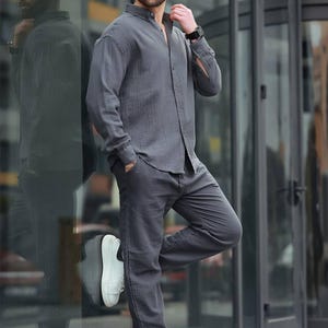May include: A charcoal grey linen shirt and matching trousers. The long-sleeved shirt is buttoned and paired with straight-leg trousers. The outfit is completed with white trainers and sunglasses. A stylish, casual ensemble.