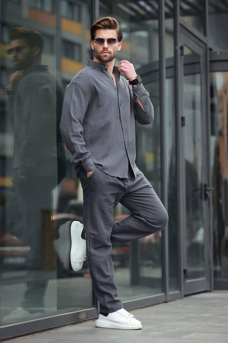 May include: A charcoal grey linen shirt and matching trousers. The long-sleeved shirt is buttoned and paired with straight-leg trousers. The outfit is completed with white trainers and sunglasses. A stylish, casual ensemble.