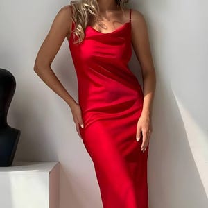 Puede incluir: Un vibrante vestido de sat&eacute;n rojo con tirantes finos y cuello desbocado. El vestido midi tiene una ca&iacute;da suave, reflejando la luz. El vestido es de color liso, con un dise&ntilde;o sencillo y elegante.