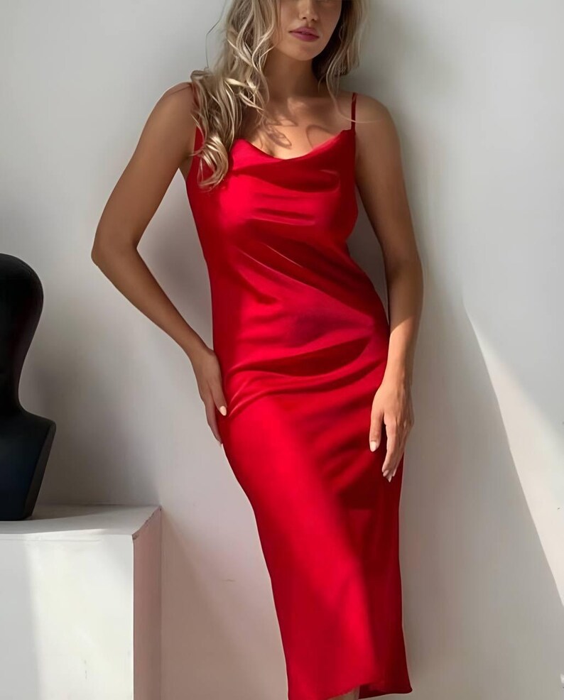 Puede incluir: Un vibrante vestido de sat&eacute;n rojo con tirantes finos y cuello desbocado. El vestido midi tiene una ca&iacute;da suave, reflejando la luz. El vestido es de color liso, con un dise&ntilde;o sencillo y elegante.