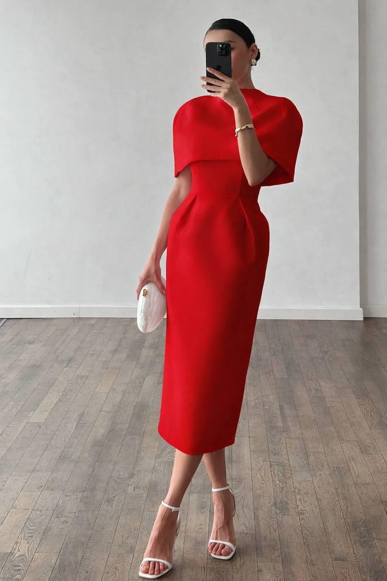 K&ouml;nnte beinhalten: Ein leuchtend rotes Midikleid mit hohem Ausschnitt und kurzen, kapuzenartigen &Auml;rmeln. Das Kleid hat ein figurbetontes Oberteil und einen geraden Rock, kombiniert mit wei&szlig;en Abs&auml;tzen und einer wei&szlig;en Clutch. Ein stilvolles Outfit f&uuml;r einen besonderen Anlass.