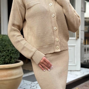 Op de afbeelding: Een beige tweedelige gebreide set. De set bestaat uit een cardigan met knopen en een klein zakje en een bijpassende knielange rok. De outfit is gemaakt van een textuur gebreide stof en heeft een ronde halslijn en lange mouwen.