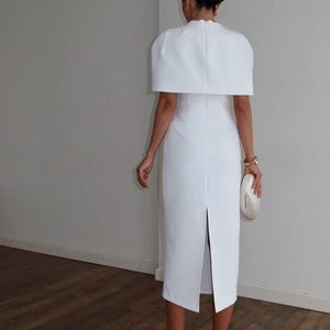 K&ouml;nnte beinhalten: Wei&szlig;es Midikleid mit capeartigem &Uuml;berwurf, hohem Ausschnitt und R&uuml;ckenschlitz. Das Kleid wird mit wei&szlig;en Riemchen-High Heels und einer perlmuttfarbenen Clutch kombiniert. Der Look ist elegant und anspruchsvoll.
