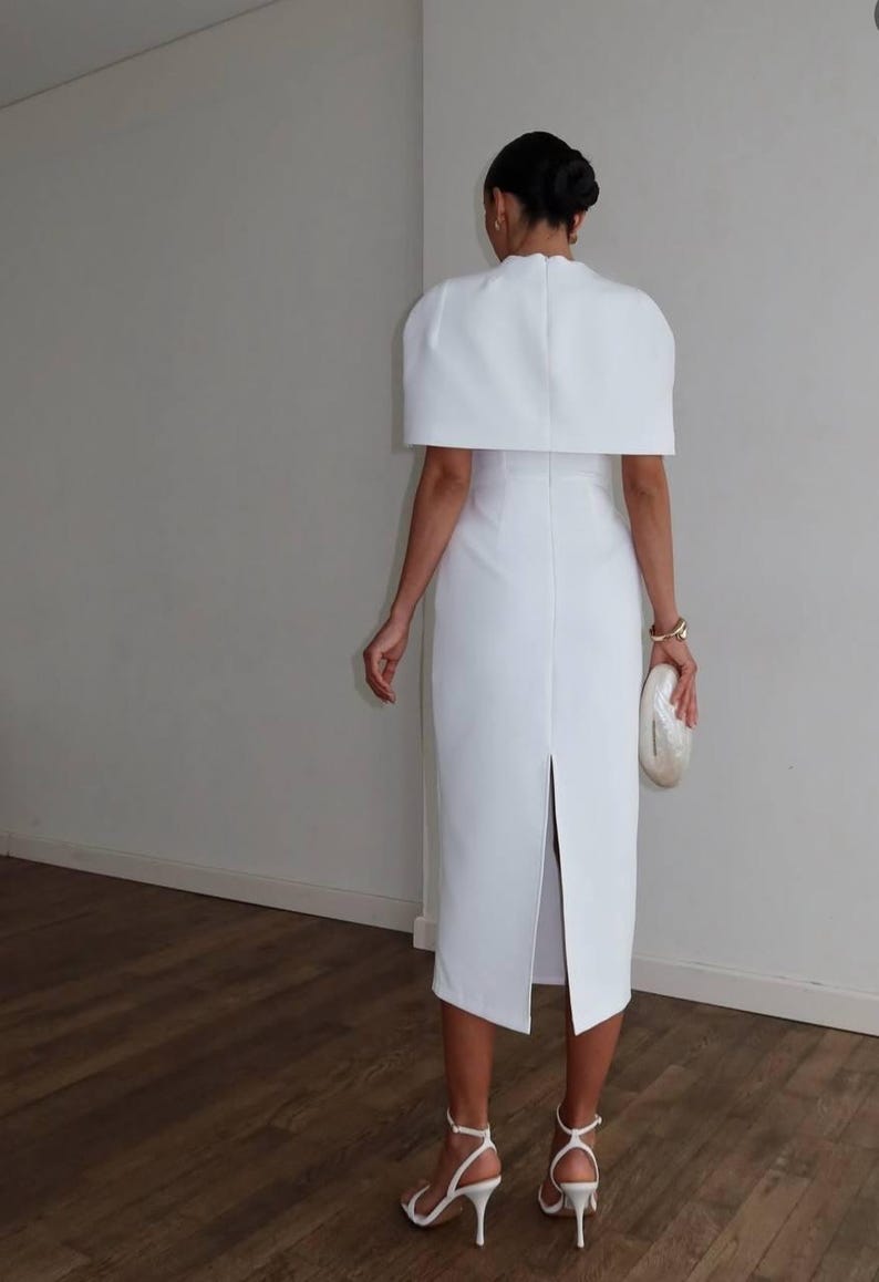 K&ouml;nnte beinhalten: Wei&szlig;es Midikleid mit capeartigem &Uuml;berwurf, hohem Ausschnitt und R&uuml;ckenschlitz. Das Kleid wird mit wei&szlig;en Riemchen-High Heels und einer perlmuttfarbenen Clutch kombiniert. Der Look ist elegant und anspruchsvoll.