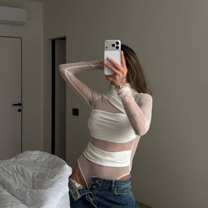 Puede incluir: Un body blanco con mangas largas y transparentes y un cuello alto. El body tiene detalles recortados y se combina con unos vaqueros azules. Se sostiene un smartphone blanco.